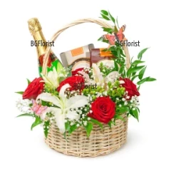 Искряща кошница, аранжирана с цветя - червени рози, бели лилиуми, свежа зеленина и подаръци - пенливо вино и 2 кутии шоколадови бонбони.